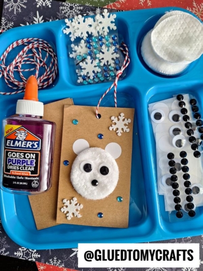 A blue tray holds craft supplies for a Winter Themed Toddler Craft: purple glue, googly eyes, black gems, snowflake stickers, twine, cotton pads, and brown cards featuring cotton pad polar bears with snowflake decorations. @GLUEDTOMYCRAFTS label below.