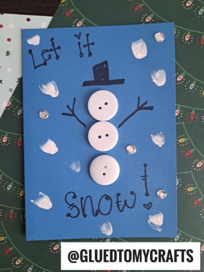 A handmade blue card features a snowman made of three white buttons, drawn stick arms, a black hat, and the words Let it snow! This Winter Themed Toddler Craft is finished with sequins, white paint dots, and @GLUEDTOMYCRAFTS at the bottom.