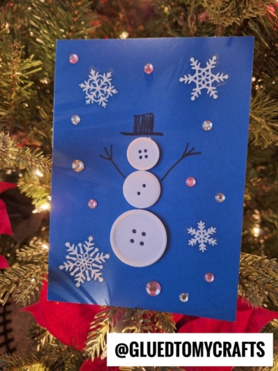 A winter themed toddler craft—this handmade holiday card features a snowman made of white buttons, drawn arms and hat, snowflake stickers, and rhinestones on blue paper, displayed in front of a decorated Christmas tree. @GLUEDTOMYCRAFTS.