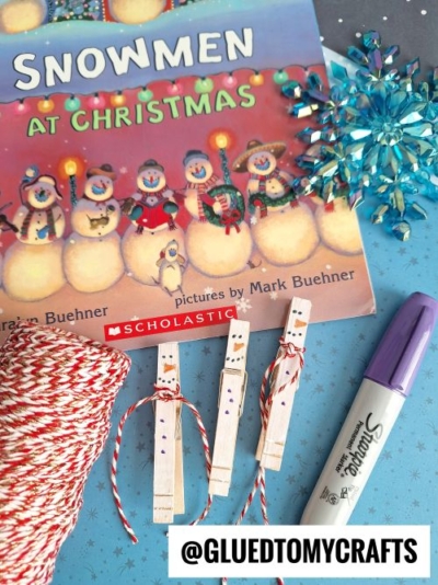 A Winter Themed Toddler Craft display features the book Snowmen at Christmas, a snowflake ornament, red and white twine, a purple Sharpie marker, and three clothespin snowman crafts on a blue, wintry background.