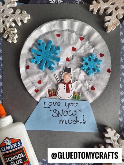 This Winter Themed Toddler Craft is a handmade snow globe featuring a snowman with a child’s photo as its head, blue foam snowflakes, small red hearts, and presents on a gray base that reads “Love you ‘snow’ much!”.