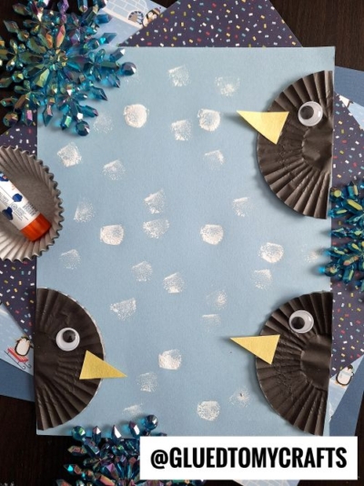A Winter Themed Toddler Craft features a blue paper with white paint spots, framed by three black paper cupcake liner owls with yellow beaks, googly eyes, shiny snowflakes, and a glue stick nearby. Text reads @GLUEDTOMYCRAFTS.