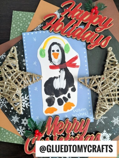 A festive handmade card, perfect as a Winter Themed Toddler Craft, features a painted penguin on white paper, surrounded by Happy Holidays and Merry Christmas signs, gold star ornaments, pine sprigs, and snowflake patterns. @GLUETOMYCRAFTS is at the bottom.