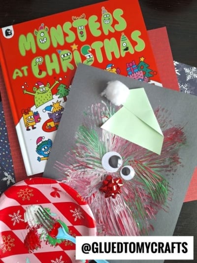 Winter Themed Toddler Craft: Children’s Christmas project featuring a green monster face made of paint, googly eyes, a cotton ball, and a paper hat, displayed on the festive book Monsters at Christmas and holiday-themed paper.