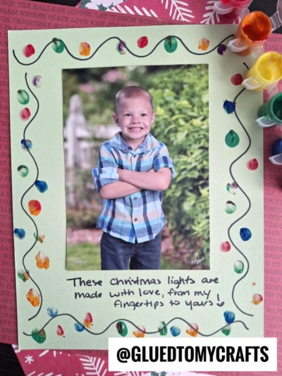 A smiling young boy in a plaid shirt poses for a photo, framed by green paper decorated with colorful painted Christmas lights and wavy lines—a charming winter themed toddler craft. Below, handwritten text says, “These Christmas lights are made with love, from my fingertips to yours!”.
