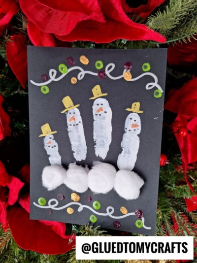 A black paper card features a Winter Themed Toddler Craft: four snowmen made from white handprints and cotton balls. Each has hats, faces, and buttons, surrounded by swirls and dots, with red poinsettias and pine branches in the background.