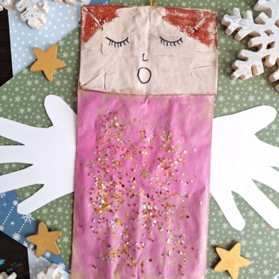 A Winter Themed Toddler Craft, this paper bag angel with a drawn face, closed eyes, and a pink glittery body rests on a green star-patterned background with white wings, wooden stars, and snowflakes. @GLUEDTOMYCRAFTS is written at the bottom.