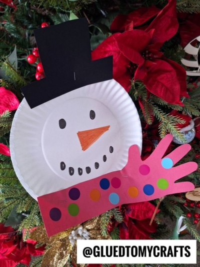 A handmade Winter Themed Toddler Craft snowman ornament, made from a paper plate with a black paper hat, orange nose, marker-drawn face, and a pink hand-shaped arm with colorful dots, hangs cheerfully on the Christmas tree.