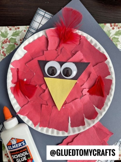 A winter themed toddler craft: a paper plate red bird made with torn red paper, a yellow paper beak, large googly eyes on black paper, and fluffy red feathers. Elmer’s glue and crafting supplies are nearby.