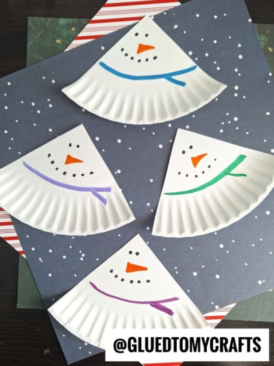 Four Winter Themed Toddler Crafts featuring paper plate snowmen with drawn faces and colorful scarves are arranged on a dark, starry background. The image has the watermark @GLUEDTOMYCRAFTS at the bottom right corner.