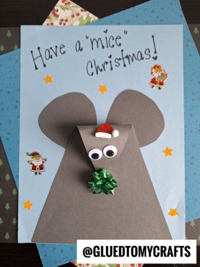 A handmade Christmas card is a charming winter themed toddler craft, featuring a mouse made from gray paper with googly eyes, a Santa hat, and bow. “Have a ‘mice’ Christmas!” is written, with festive star and Santa stickers decorating the card.