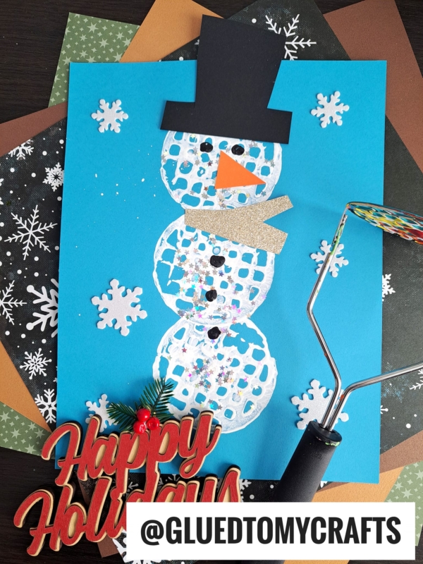 A Winter Themed Toddler Craft: A snowman made from painted potato masher prints is on blue paper, decorated with a black hat, orange nose, gold scarf, snowflakes, and glitter. Craft papers and a Happy Holidays decoration are nearby.