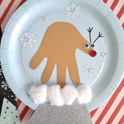 A Winter Themed Toddler Craft featuring a paper reindeer made from a brown handprint on a blue plate, complete with googly eyes, a red nose, snowflakes, cotton ball snow, and sparkly silver paper at the bottom.