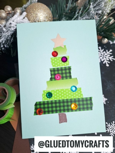 A handmade Winter Themed Toddler Craft Christmas card features a tree made of layered green and plaid washi tape, colorful gems as ornaments, and a star on top. Pine branches and gold ornaments are in the background. @GLUEDTOMYCRAFTS is written at the bottom.