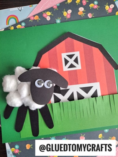 A handprint sheep made with cotton balls and googly eyes stands in front of a red barn, with green paper grass in the foreground. The @GLUEDTOMYCRAFTS watermark is at the bottom right.