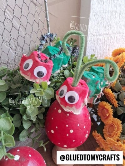 Two egg carton hungry caterpillars made from painted egg cartons with googly eyes and pipe cleaner antennae sit on a red mushroom decoration, surrounded by green plants and yellow flowers.