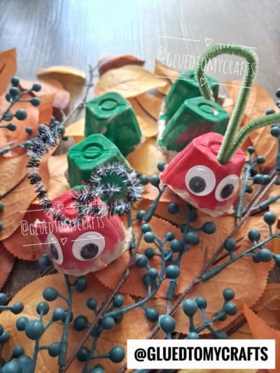Egg carton caterpillars and egg carton hungry caterpillar crafts with googly eyes and pipe cleaner antennae sit on fake autumn leaves and berry branches. Craft by @GLUEDTOMYCRAFTS.