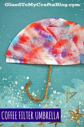 A colorful coffee filter umbrella painted with red, blue, and purple sits on teal paper as an umbrella, with a gold pipe cleaner as the handle; text reads “COFFEE FILTER UMBRELLA”—a fun umbrella craft for kids.
