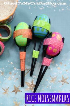 Three colorful fiesta noisemakers, crafted from plastic spoons and decorated plastic egg noisemakers filled with rice, are wrapped in patterned tapes and displayed on a blue star-patterned surface. Text: RICE NOISEMAKERS.