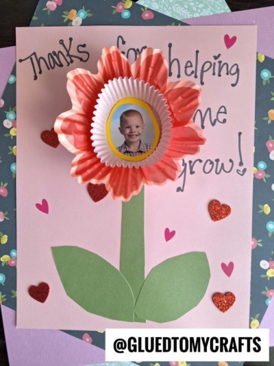 A handmade card features a cupcake liner flower with a child’s photo in the center, green paper stem and leaves, red glitter hearts, and handwritten text: “Thanks for helping me grow!” on a pink background.