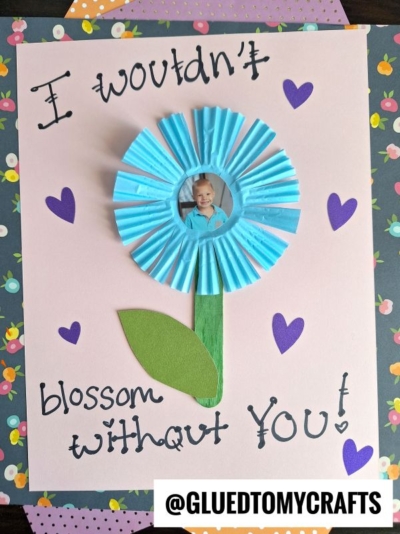 A handmade card features a blue cupcake liner flower with a child’s photo in the center, green paper leaf and stem, purple hearts, and the handwritten words “I wouldn’t blossom without you!” on a pink background.