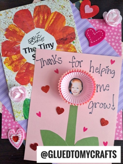 A handmade card with a child’s photo in a cupcake liner flower, reading “Thanks for helping me grow!” sits next to The Tiny Seed book. Pink hearts, paper flowers, and a striped background add a sweet touch.
