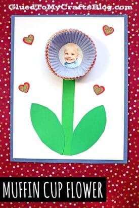 A craft project featuring a cupcake liner flower made with a child's photo in the center, green paper stem and leaves, and heart decorations on a white background with a red polka dot border.