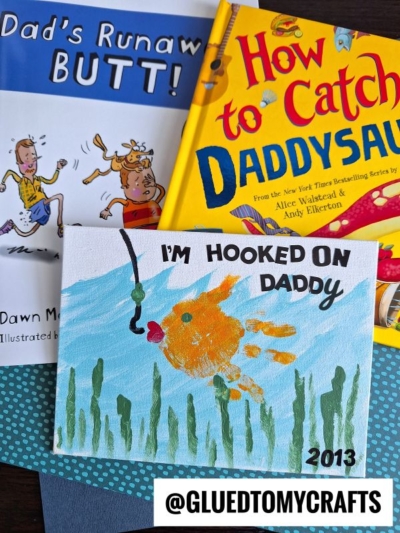 Three items: two children's books, Dads Runaway Butt! and How to Catch a Daddysaurus, plus an "I'm hooked on daddy canvas" featuring a handprint goldfish and the text “I’M HOOKED ON DADDY 2013.”.