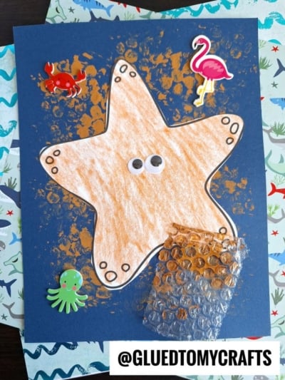 A children’s craft featuring a large orange bubble wrap print starfish with googly eyes on blue paper, decorated with crab, octopus, and flamingo stickers, ocean-themed paper in the background. The handle @GLUEDTOMYCRAFTS is at the bottom.