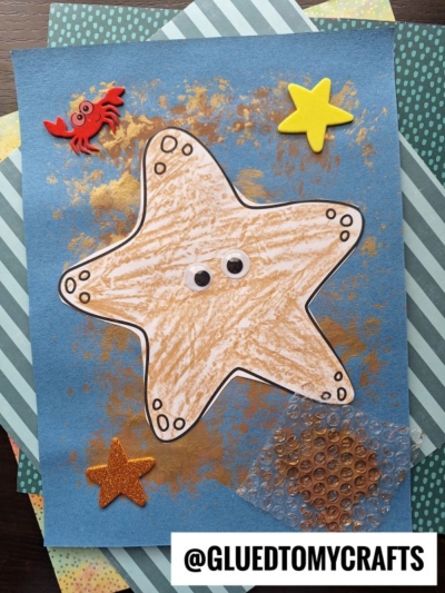 A handmade bubble wrap print starfish craft on blue paper with googly eyes, gold paint, a fake crab, two star stickers, and patterned papers underneath. Text reads @GLUEDTOMYCRAFTS at the bottom.