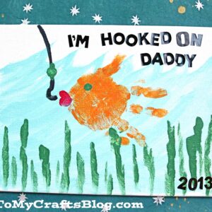 A child’s handprint painted as an orange fish swims toward a fishing hook on this “I’M HOOKED ON DADDY” canvas card. Blue, green, and white with star patterns make it special. GluedToMyCraftsBlog.com at the bottom.