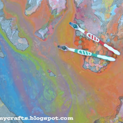 Two toothbrushes with colored handles lie on a concrete surface covered in a mix of bright, swirling liquid chalk and paint splatters in orange, blue, pink, and green hues. Text Gluedtomycrafts.blogspot.com appears at the bottom.