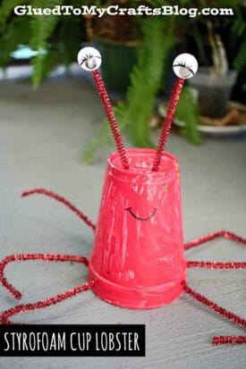 A styrofoam cup lobster, painted red and decorated with pipe cleaner legs, antennae, googly eyes, and a drawn-on smile. In the background are a plant and a craft blog watermark.