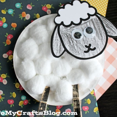 A charming cotton ball sheep craft made with a paper plate, a drawn face and ears, googly eyes, and stick legs sits on colorful floral and plaid paper backgrounds.