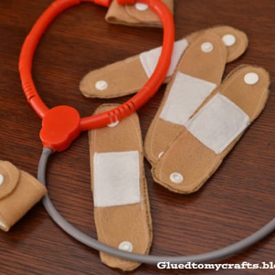 A toy stethoscope and several DIY band-aids, including handmade felt bandages and a cuff in tan, white, and red, are arranged on a wooden surface.