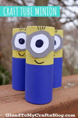 Three craft tube Minions made from painted cardboard tube minion bodies with paper eyes, mouths, and hair stand on a wooden surface. A label above reads Craft Tube Minion. Trees are visible in the blurry background.
