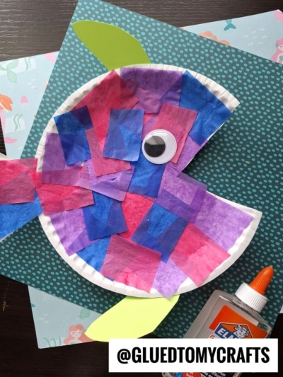 A colorful paper plate fish craft features pink, purple, and blue tissue squares with a googly eye and a blue paper tail. Perfect for creating your own tropical paper fish! Elmers glue sits nearby on a patterned background. Text reads @GLUEDTOMYCRAFTS.
