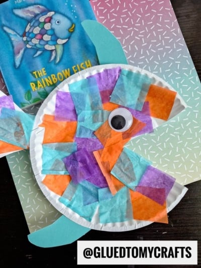 A vibrant paper plate fish craft, inspired by tropical paper fish, is adorned with colorful tissue squares and a googly eye. It rests beside The Rainbow Fish book on a patterned background. Username @GLUEDTOMYCRAFTS is displayed below.