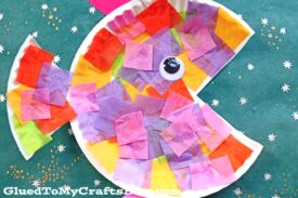 A vibrant tropical paper fish crafted from a paper plate, adorned with tissue paper squares and a googly eye, set against a green background dotted with white, star-like spots.