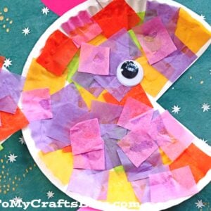 A vibrant tropical paper fish crafted from a paper plate, adorned with tissue paper squares and a googly eye, set against a green background dotted with white, star-like spots.