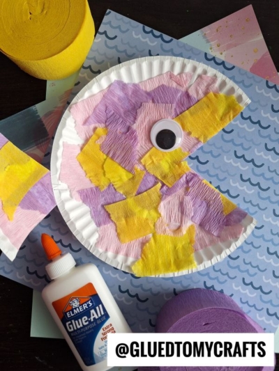 A paper plate fish decorated with yellow, purple, and pink tissue paper to look like Pac-Man with a large googly eye, on top of patterned paper. Elmer’s glue and rolls of crepe paper are nearby.