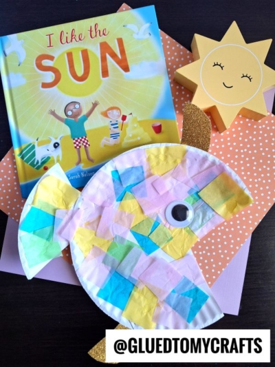 A colorful tropical paper fish craft sits on pink and orange polka-dot paper, next to a smiling sun decor and the book I Like the Sun. The handle @GLUEDTOMYCRAFTS appears at the bottom.