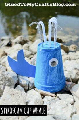 A styrofoam cup whale painted blue, complete with a paper tail, googly eye, and white pipe cleaner water spouts, sits on rocks by a pond. Text reads “GluedToMyCraftsBlog.com” and “Styrofoam Cup Whale.”.