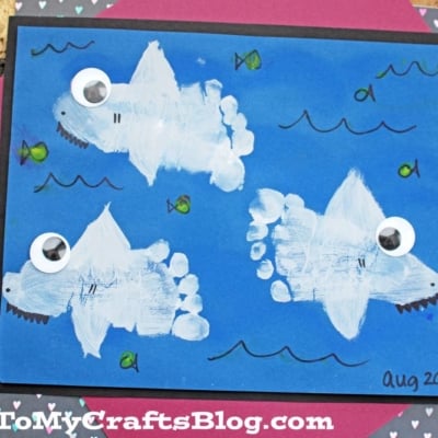 A children’s craft featuring three footprint sharks painted with white footprints on blue paper, each with a googly eye. Fish, waves, and details are drawn with markers. The piece is signed Aug 2013.