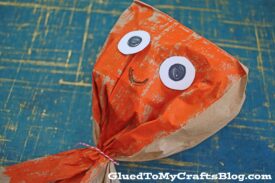 A homemade craft jellyfish with an orange-painted paper body, large paper eyes, a drawn smile, and tied at the bottom with red and white string sits on a blue textured surface beside a playful paper bag octopus.