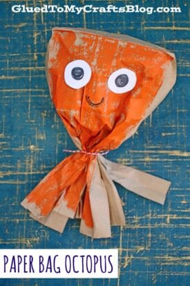 A paper bag octopus painted orange with white googly eyes, a drawn-on smile, and tied with a string to form tentacles sits on a blue and yellow textured background.