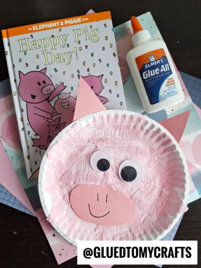 A children’s book titled Happy Pig Day! sits beside a paper plate pig craft made with pink paper and googly eyes. Elmer’s Glue and assorted craft papers complete the scene. @GLUEDTOMYCRAFTS is written at the bottom.