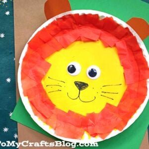 A paper plate lion craft featuring a yellow center, red and orange tissue paper mane, googly eyes, black nose, drawn whiskers, and brown paper ears sits on a green and brown background.
