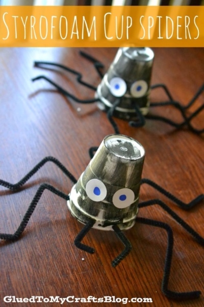 Two black-painted Styrofoam cups turned upside down with googly eyes and black pipe cleaner legs, resembling a styrofoam cup spider, rest on a wooden surface. Text reads STYROFOAM CUP SPIDERS.