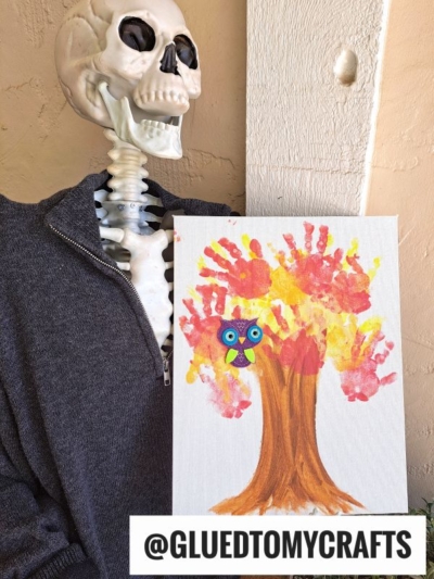 A skeleton in a gray sweater holds a painting of a handprint fall tree made from colorful handprints and a small owl. Text at the bottom reads @GLUEDTOMYCRAFTS.